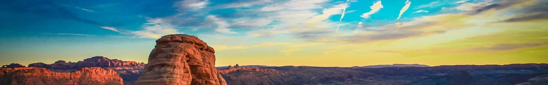 A wide panoramic desert scene with a warm red rock arch in the foreground, set against expansive canyon terrain under a vivid blue-to-gold sunset sky.
