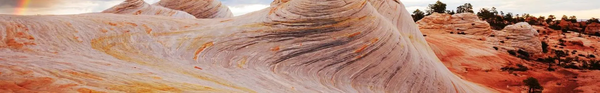 A dramatic rainbow arcs across a cloudy sky above layered, swirling sandstone formations in warm cream, orange, and red tones, with the landscape filling the foreground.