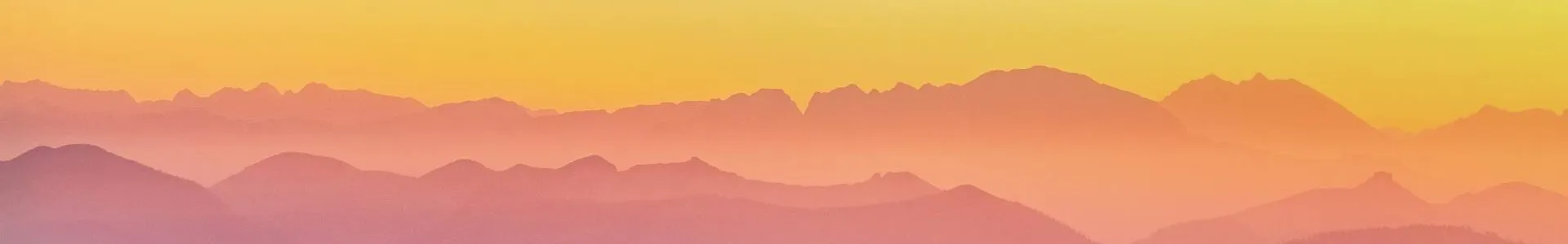Soft pastel mountain ridges fade into the distance in layered silhouettes of purple, pink, and orange beneath a pale yellow sky, with a small lone hiker standing on a dark foreground ridge.